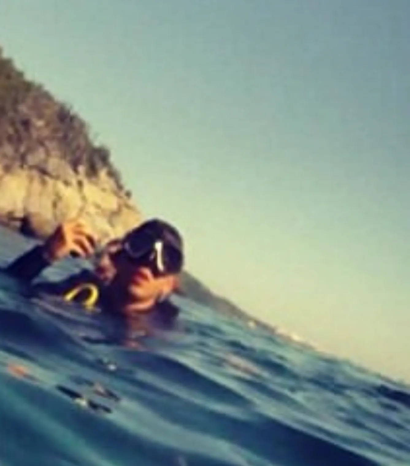 Raphael at the surface wearing a dive mask with a rocky coast behind