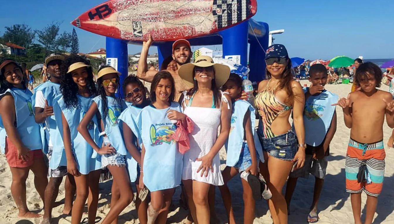 Kids in light blue vests on the beach with adults, in front of an inflatable arch with a surfboard on top