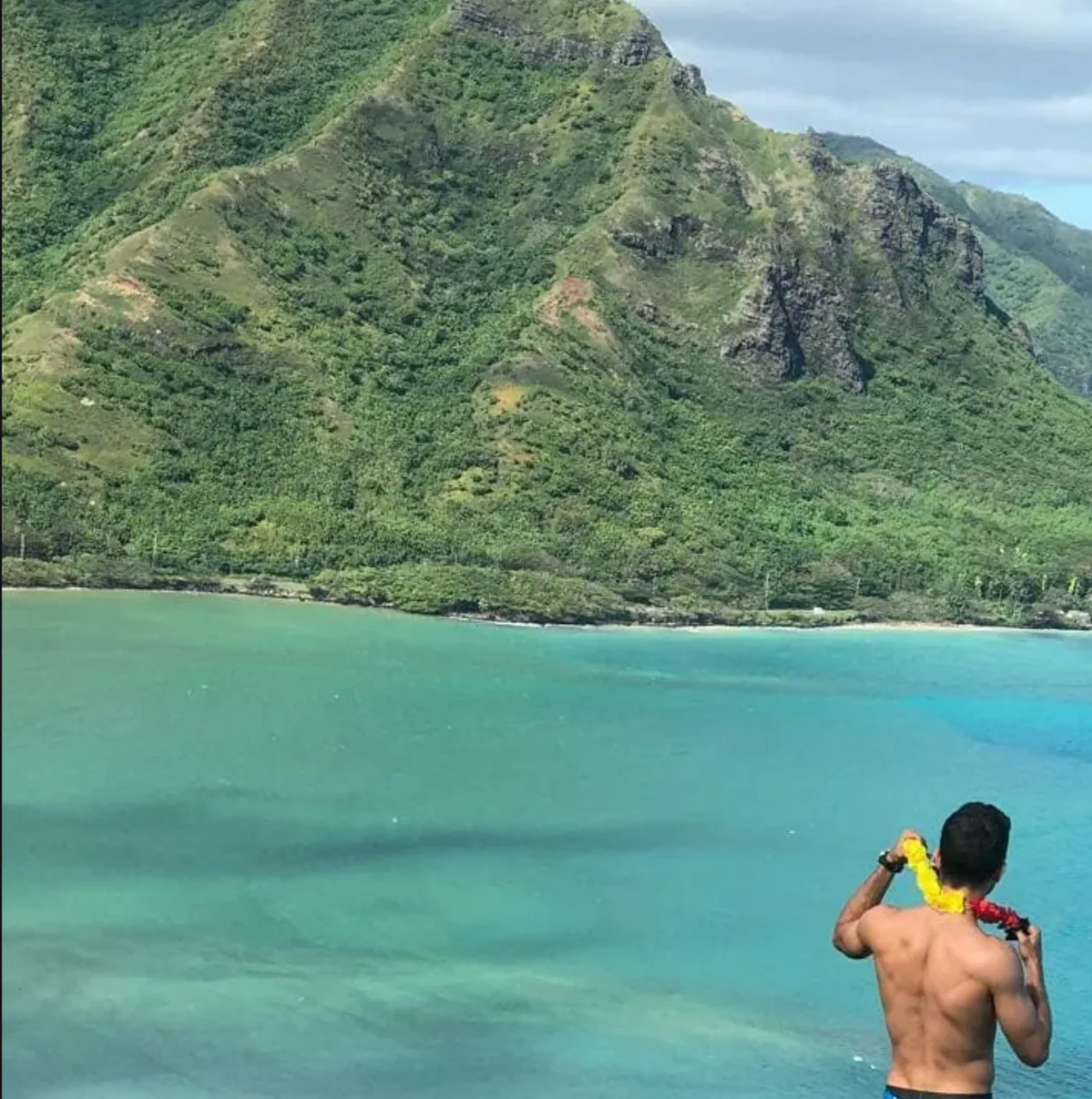 Raphael from behind with a flower lei looking at a green mountain and turquoise bay