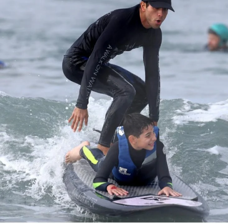 Raphael in a wetsuit helping a kid ride a wave on a surfboard