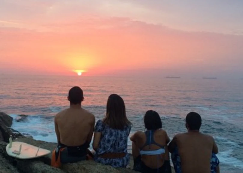 Friends sitting on the rocks watching the sunset with a surfboard beside them