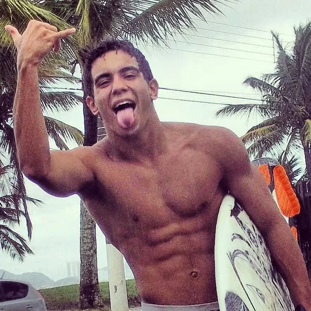Raphael with a shaka sign and a surfboard under palm trees